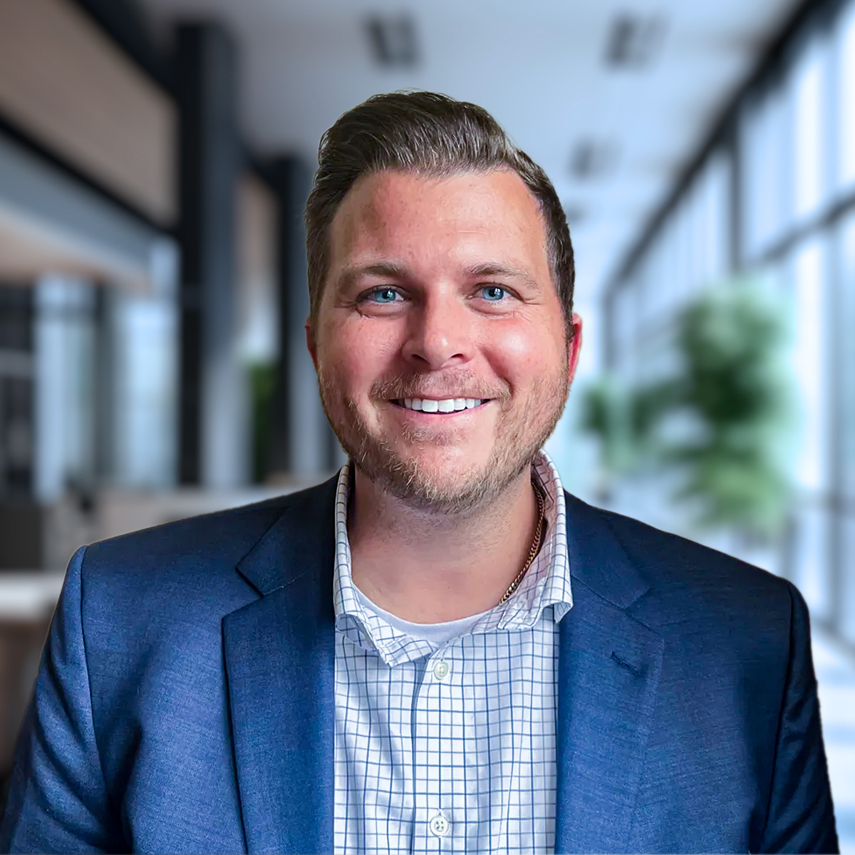 A smiling man in a blue blazer and checked shirt stands in a modern, bright office hallway with large windows and blurred greenery in the background.