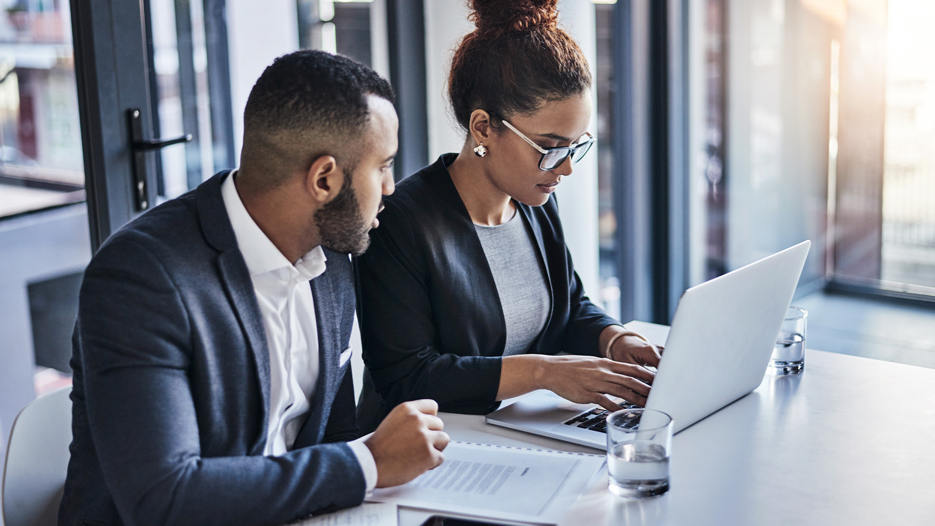 Two professionals sit at a desk in an office, reviewing a dental employment contract. The woman types on a laptop while the man looks on, both dressed in business attire. Papers and glasses of water are on the table by a large window.