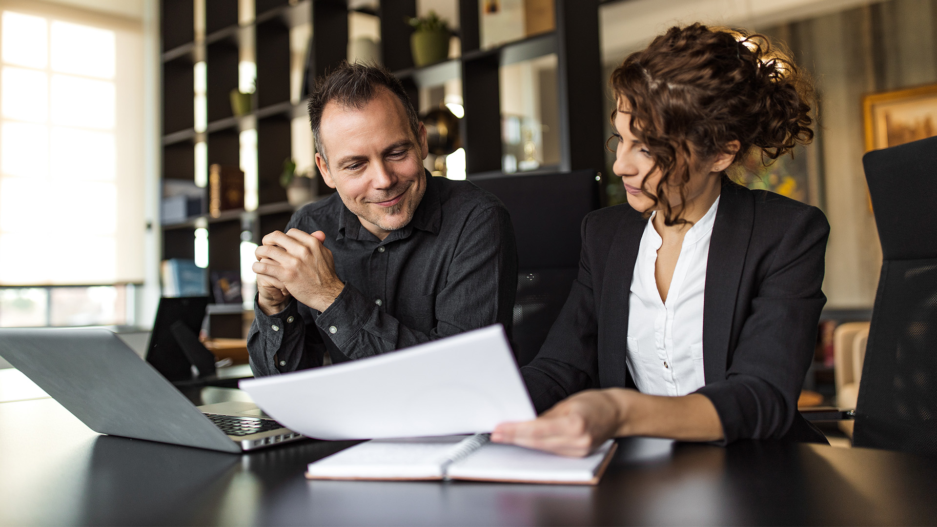 Two colleagues sit at a desk in an office. The woman, a dental service organization attorney, shows documents to the man, who is smiling. A laptop and notebooks are on the table, with bookshelves in the background.
