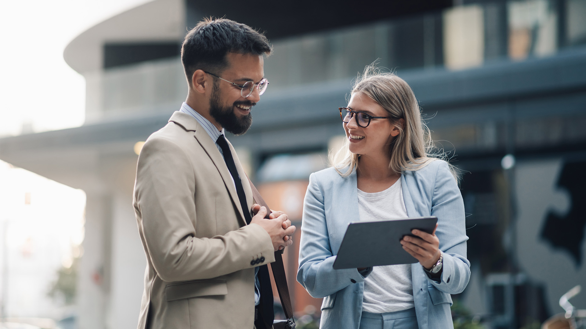 Two business professionals, a man and a woman, stand outdoors in suits and glasses. The woman, possibly consulting a dental entity formation attorney St. Louis, smiles as she discusses something on her tablet with the man in a friendly manner.
