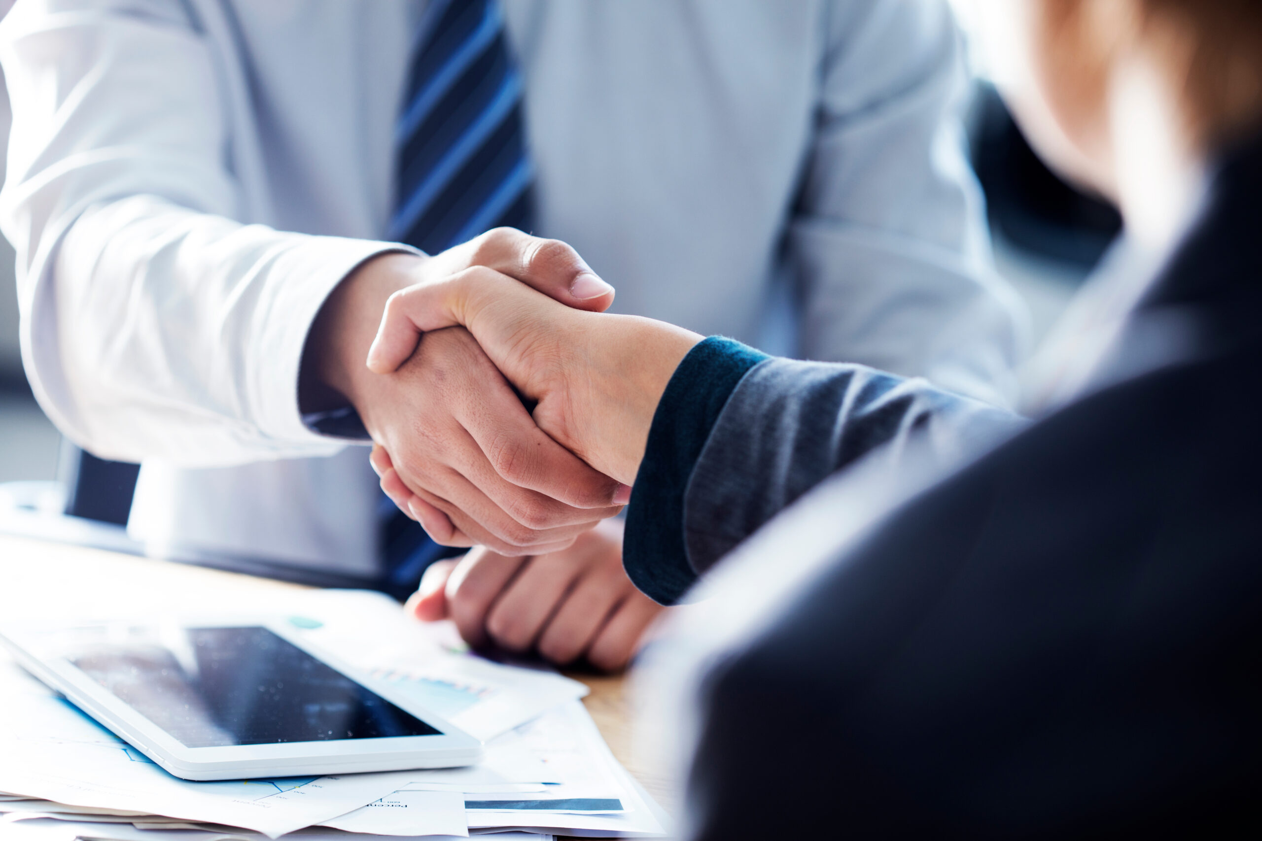 Two people in business attire shaking hands over a desk with papers and a tablet, symbolizing agreement or partnership. The focus is on the handshake, with faces out of view.