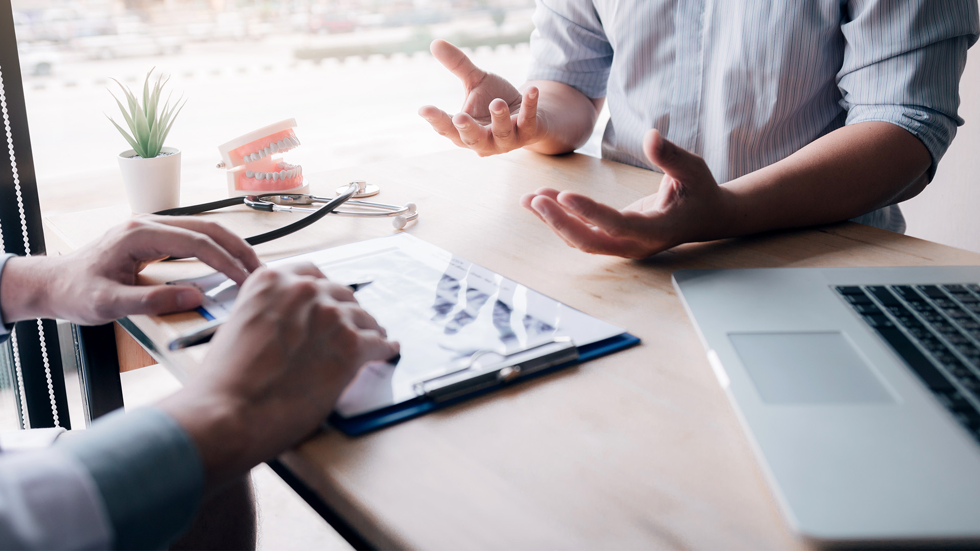 Two people sit at a desk discussing dental X-rays. One points at the X-rays on a clipboard while the other gestures. A laptop, dental model, stethoscope, and notes from a dental mergers and acquisitions lawyer are on the table.