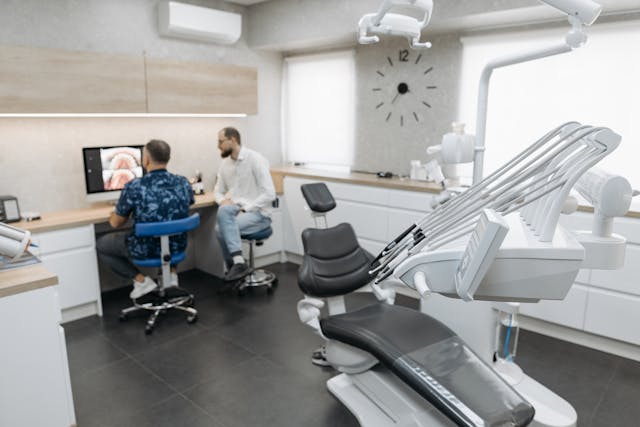A modern dental office with two men sitting at a desk reviewing dental images on a computer; dental equipment and an empty examination chair are in the foreground.