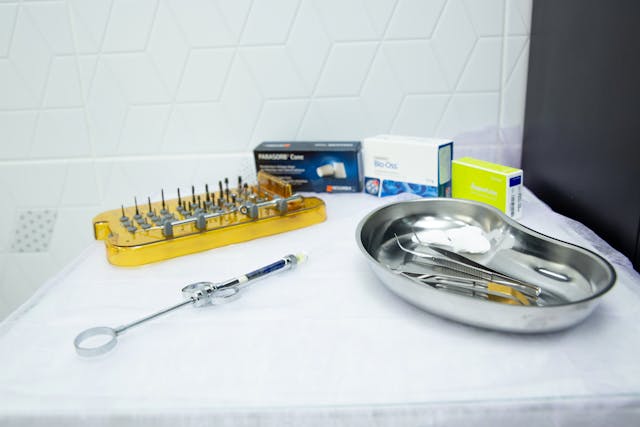 Dental instruments, including a surgical tray with tools, a gold-colored drill bit holder, and a syringe, are arranged on a white surface.